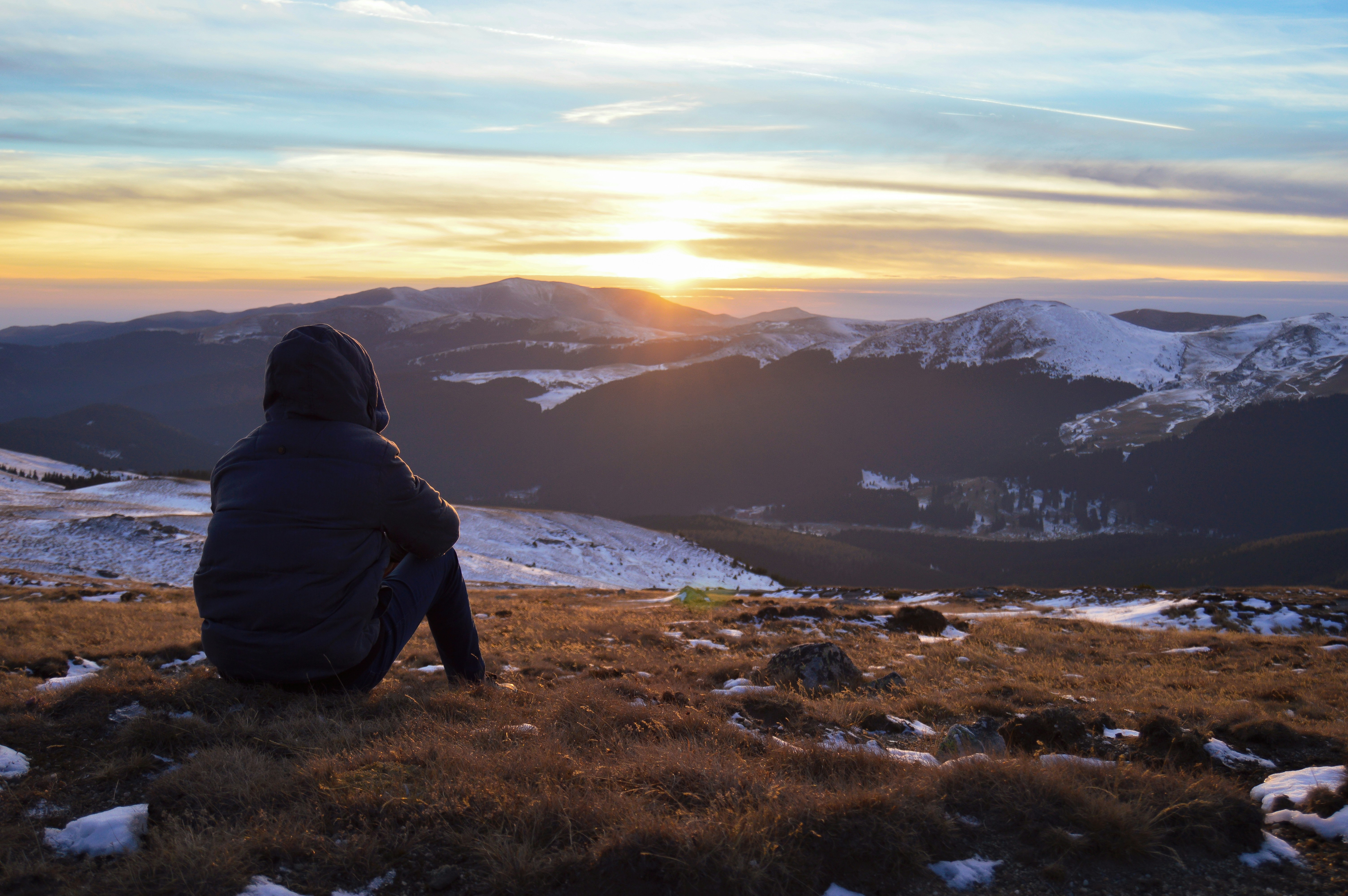 A person in a black hooded jacked sits on top of a mountain and looks out over water and mountains at the sun rising.
